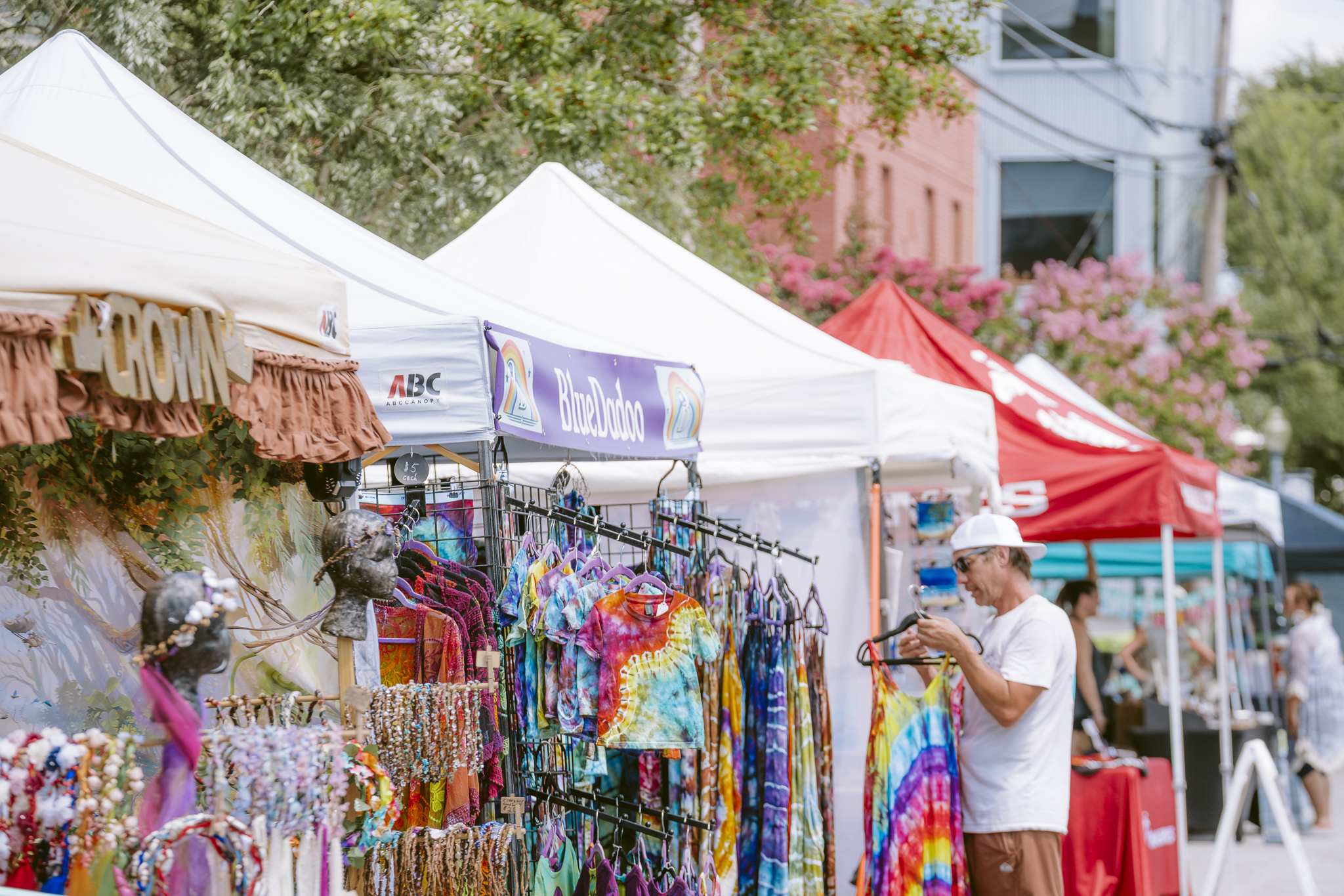 Colorful vendor booths at an outdoor festival marketplace, featuring tie-dye clothing and handmade jewelry for sale, with a shopper browsing items under white canopy tents on a sunny day
